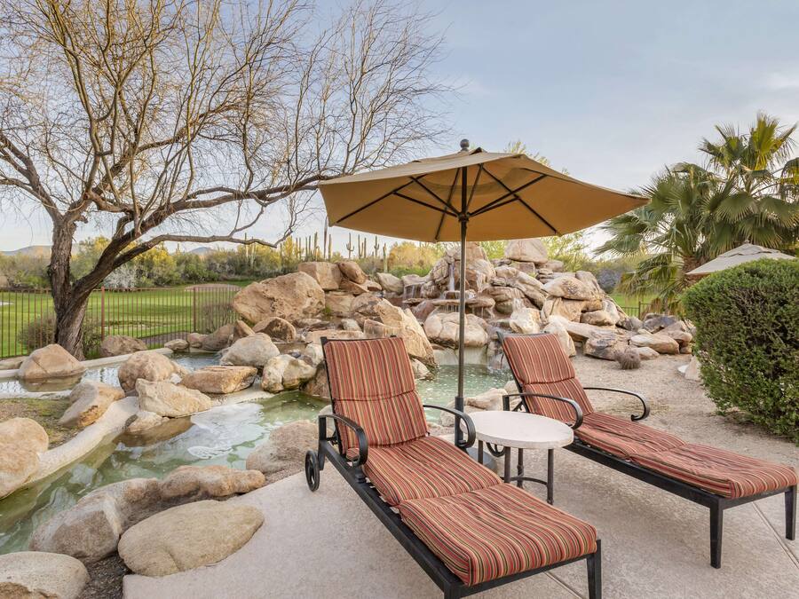 A patio with two lounge chairs and a table, a large umbrella, and a small water feature surrounded by rocks.