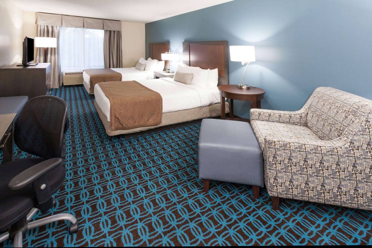 A hotel room with two beds, a desk with a lamp, a chair, and a patterned carpet.