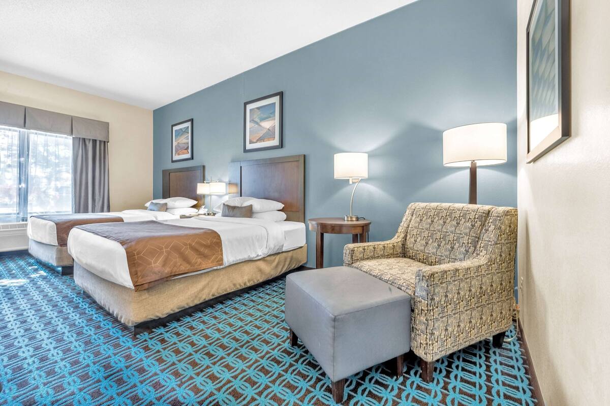 A hotel room with two beds, a chair, an ottoman, a small table, and a lamp.