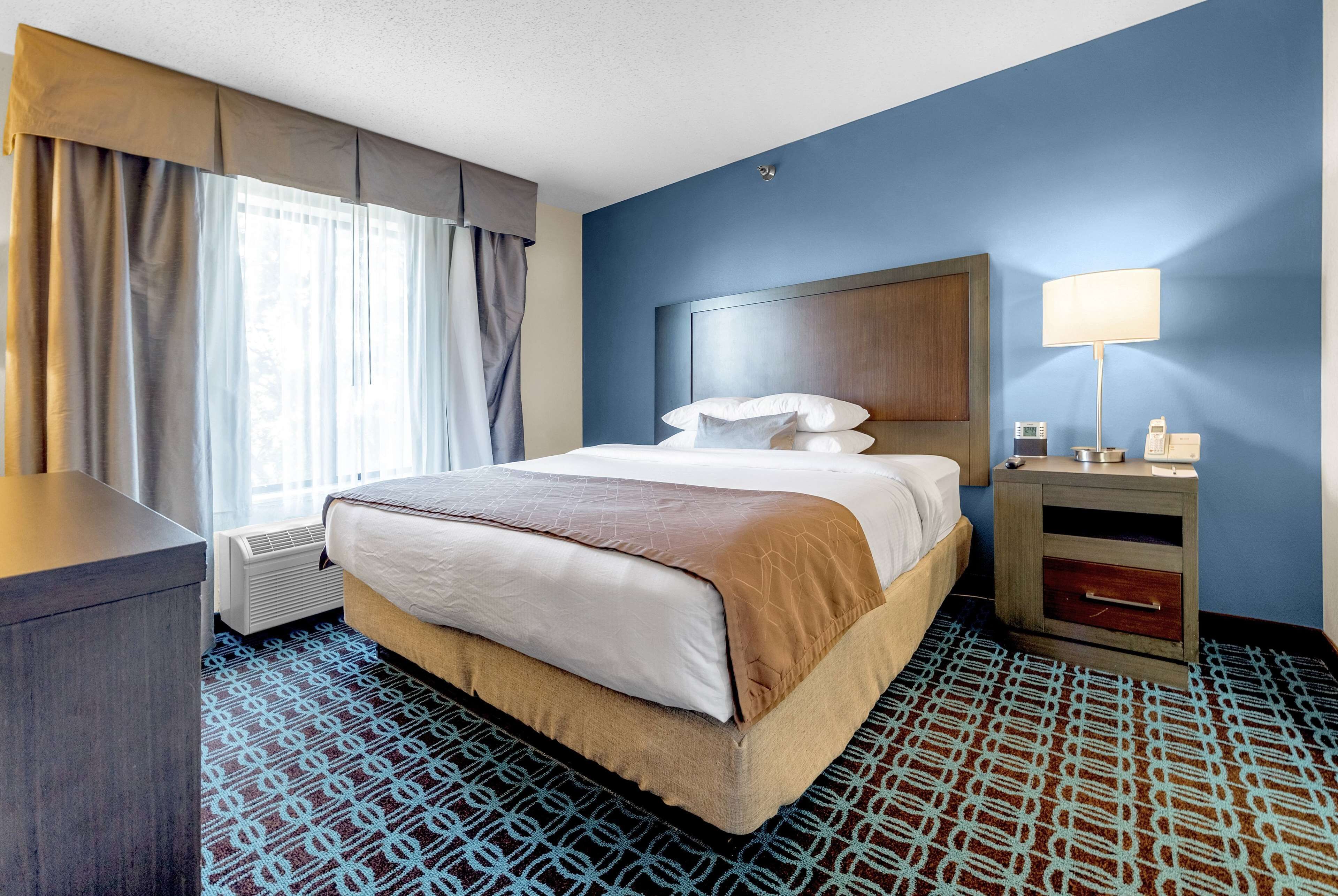 A hotel room with a bed, bedside table, lamp, and air conditioning unit.