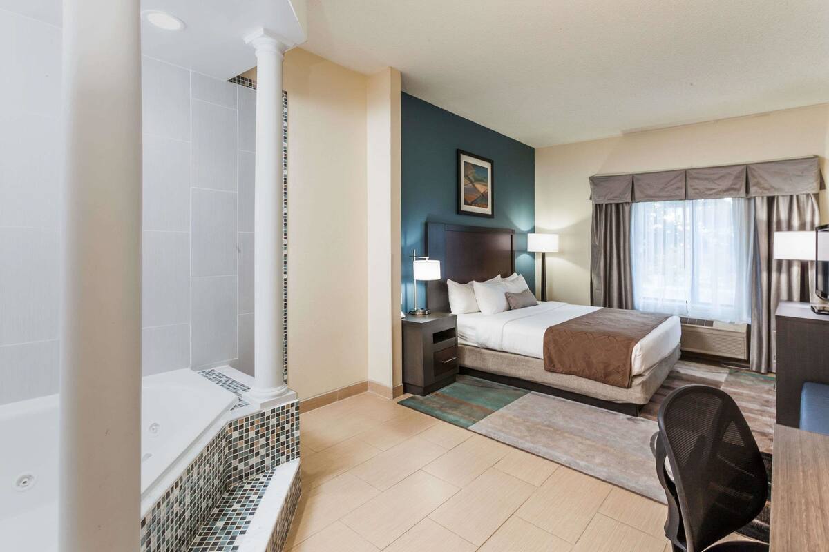 A hotel room with a bed, bedside tables, a desk, a chair, a bathtub, and a window with curtains.