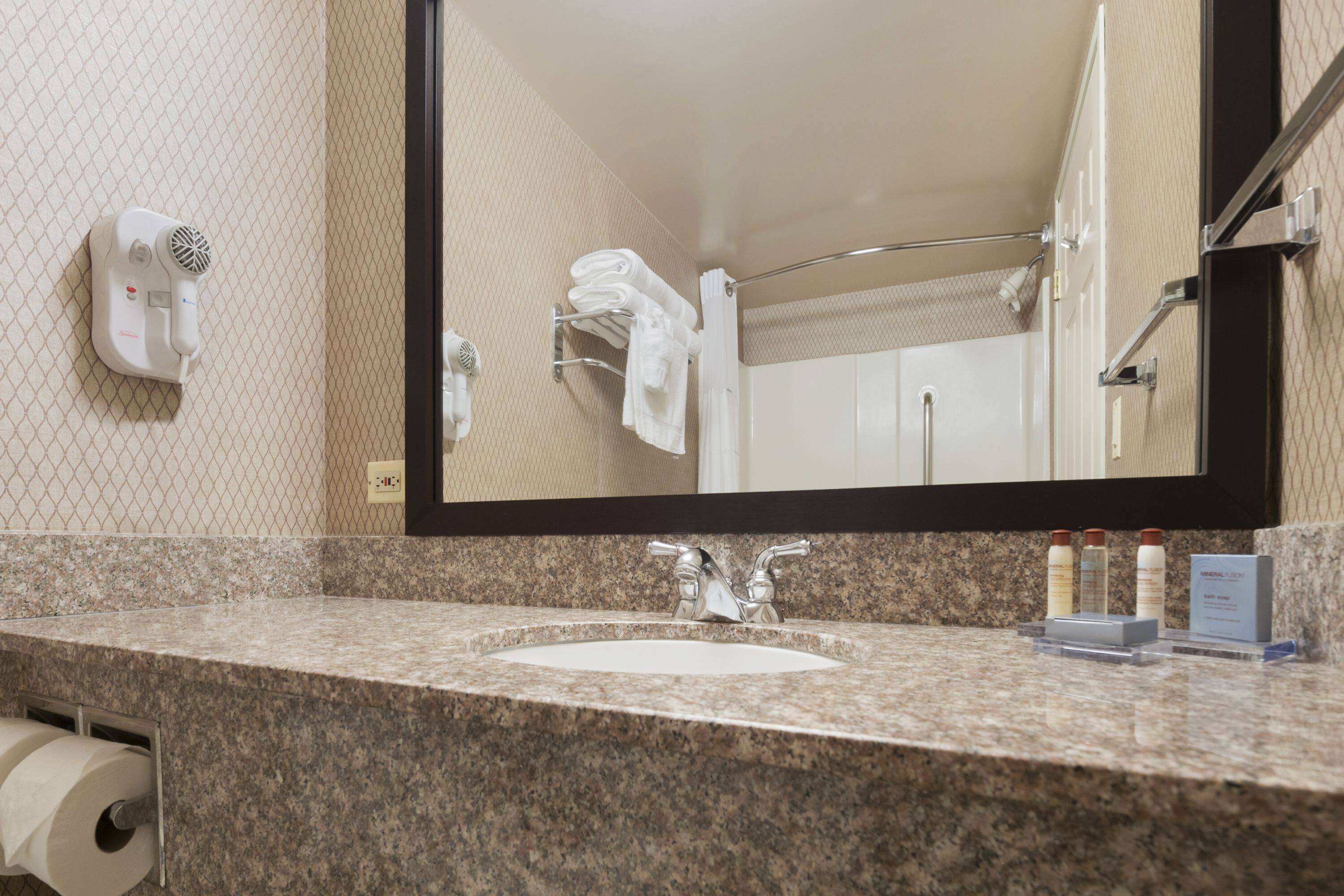 A hotel bathroom with a granite countertop, a sink, a mirror, a towel rack, and a hairdryer.