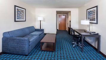 Une chambre d’hôtel avec un tapis à motifs bleus, un canapé bleu, une petite table en bois, un bureau avec une lampe et deux œuvres d’art encadrées accrochées aux murs.