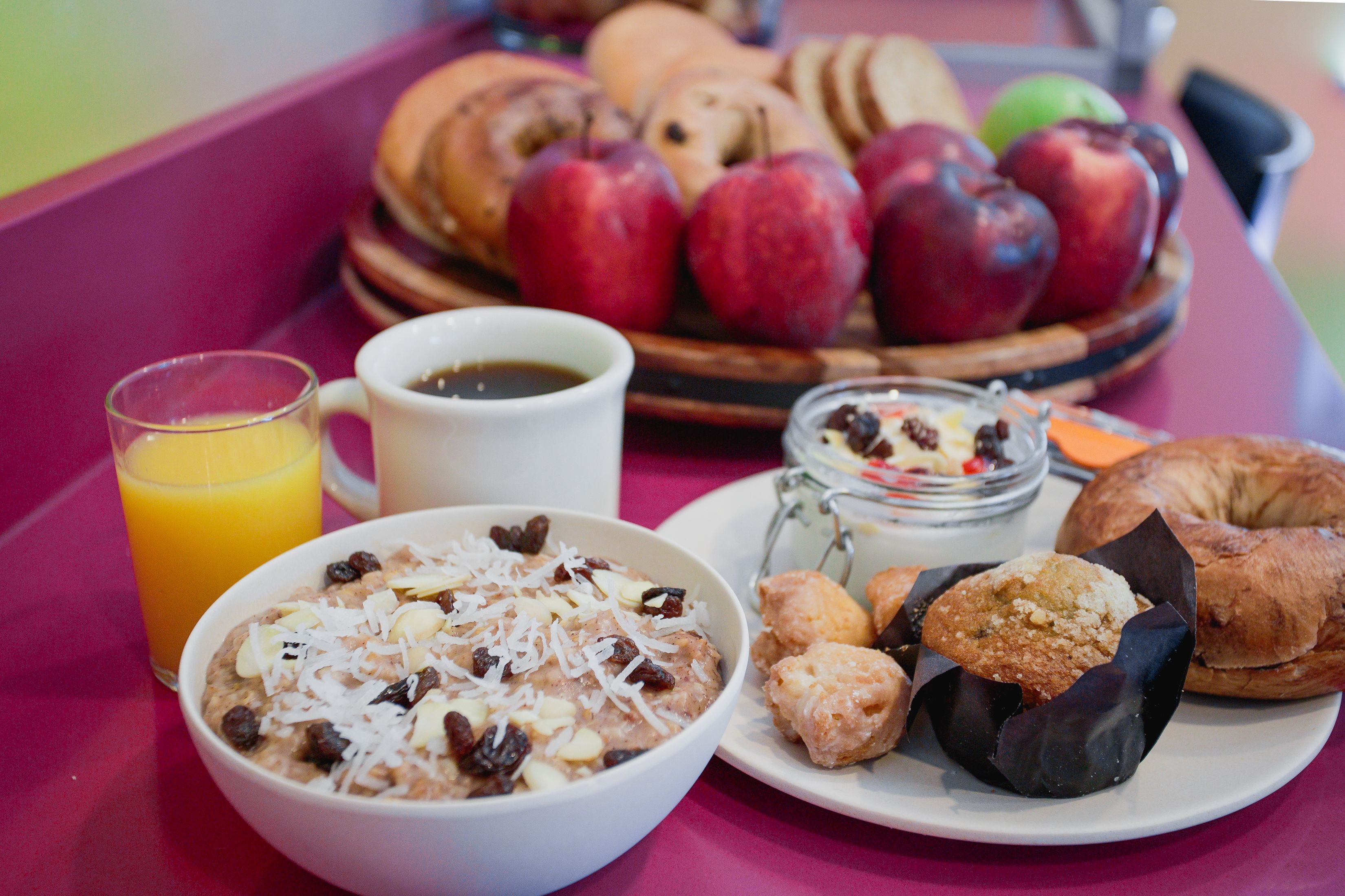Un plateau de petit-déjeuner avec un bol de céréales, un verre de jus d’orange, une tasse de café, une assiette de viennoiseries et un panier de pommes.