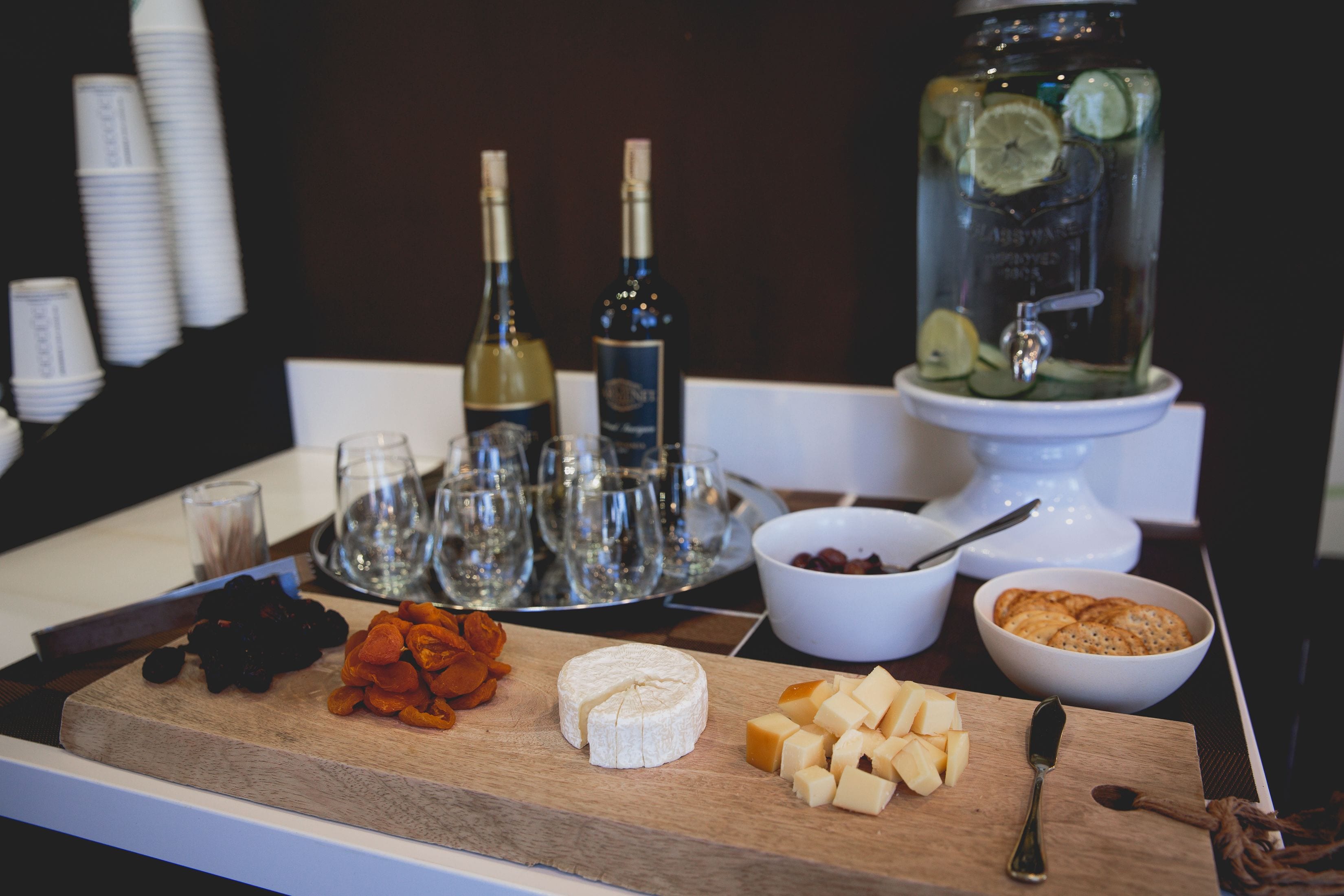 A cheese board with various cheeses, a jar of pickles, and a bowl of nuts on a wooden board.