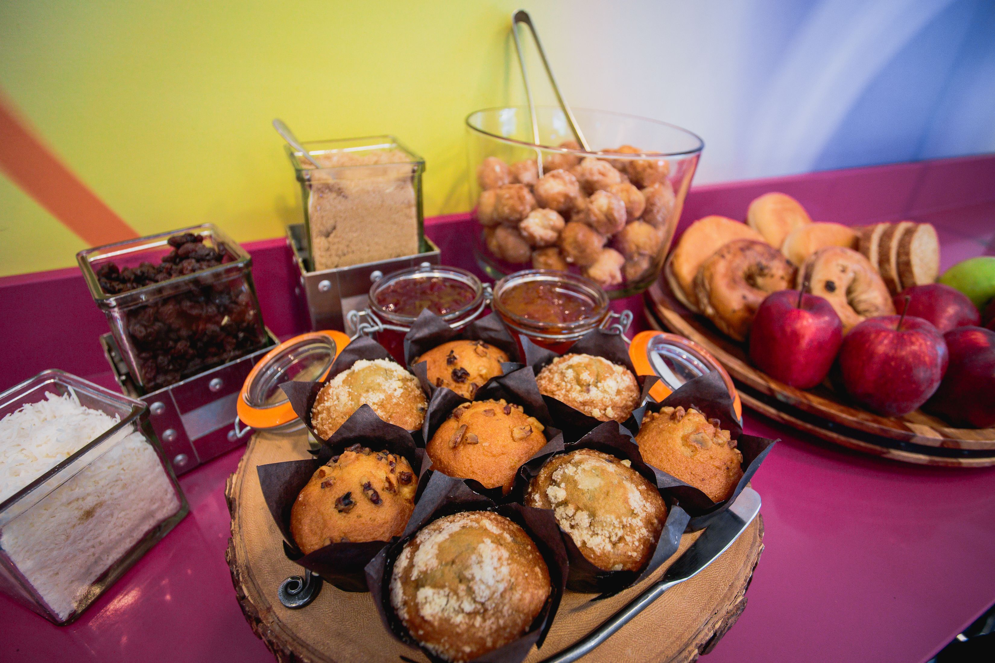 A table with muffins, a bowl of apples, and various jars of spreads.