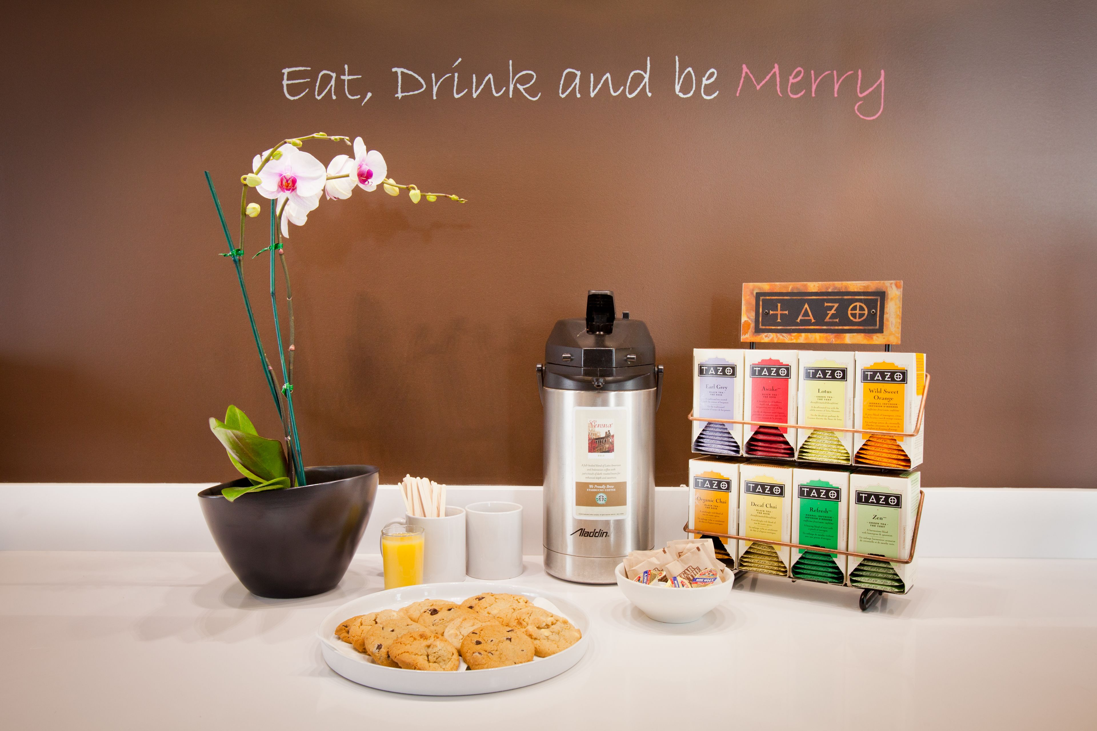 A breakfast setup with a coffee maker, a tray of cookies, a bowl of fruit, and a sign with the word "TAZO" on a cart.
