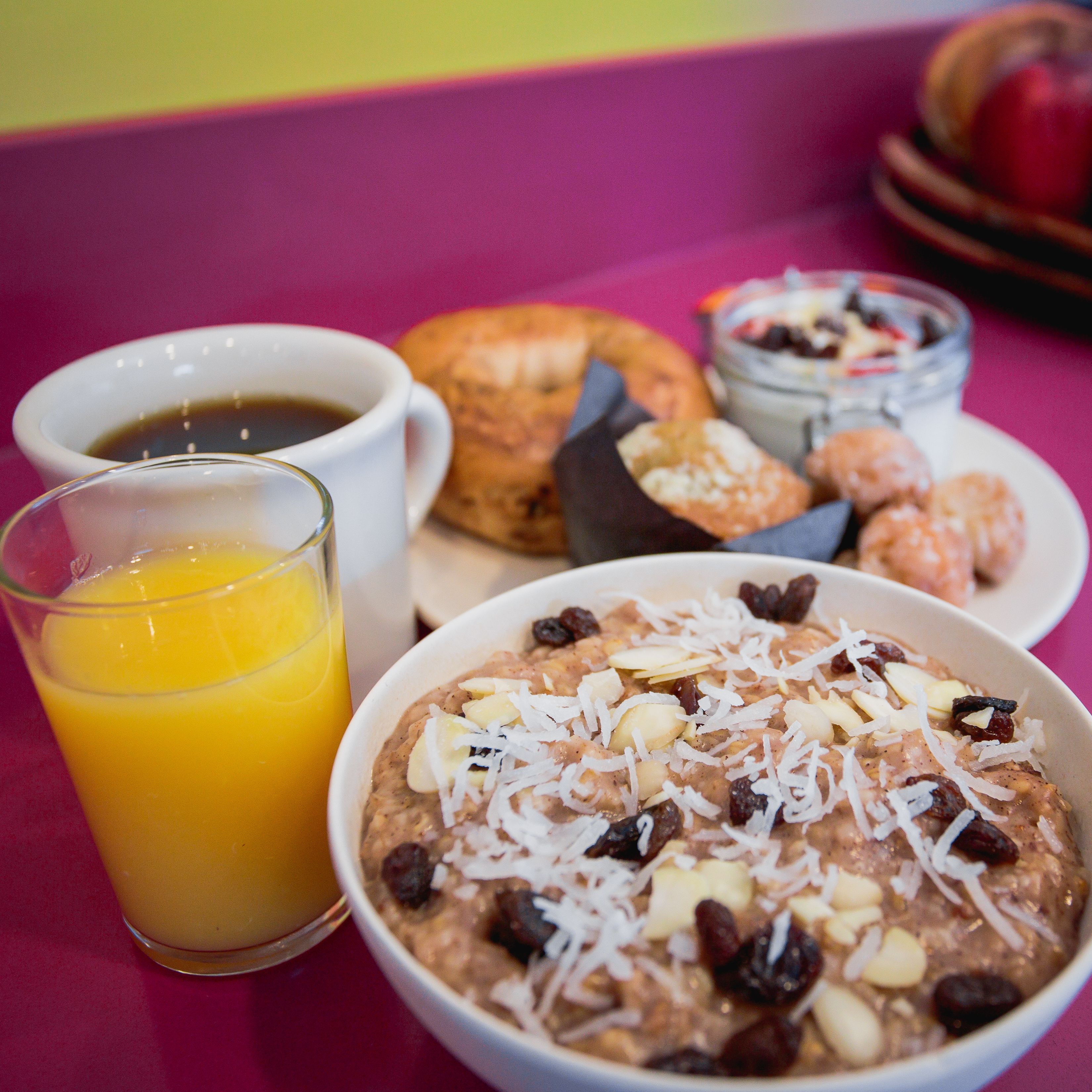 Un desayuno con un tazón de avena cubierto de nueces y pasas, un vaso de jugo de naranja, una taza de café, un croissant y un pequeño tazón de yogur con bayas.
