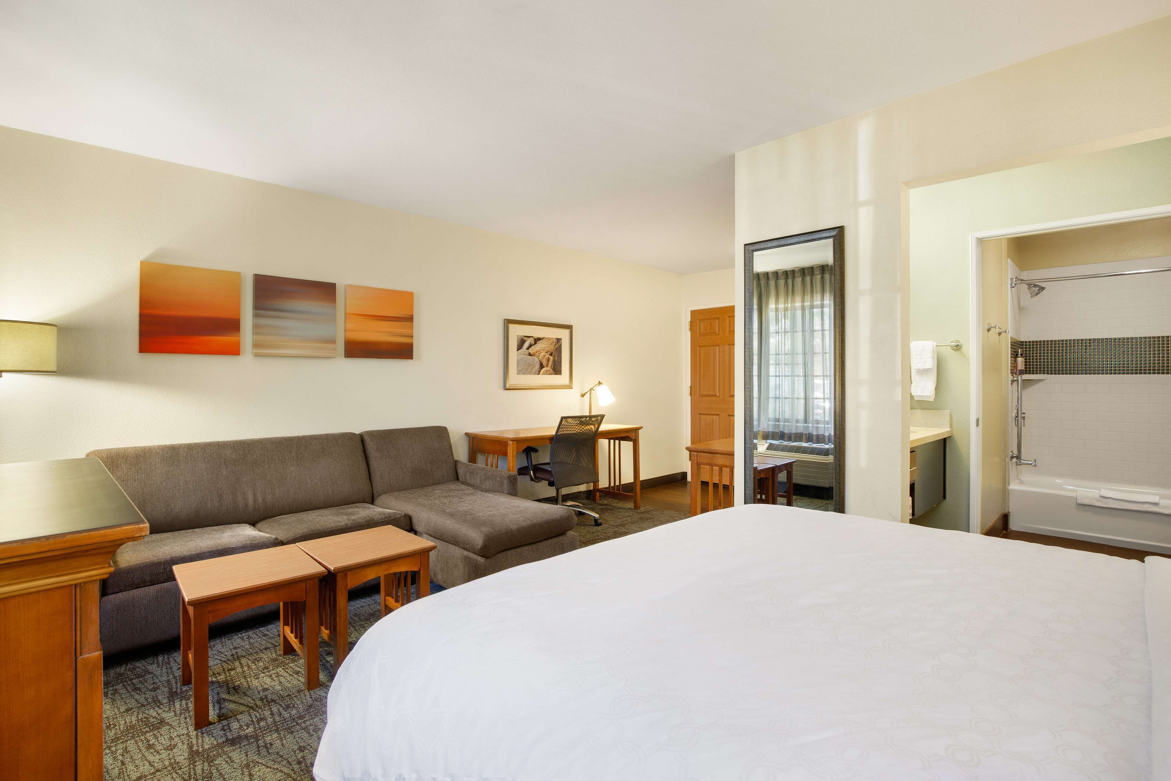 Une chambre d’hôtel avec un grand lit, un canapé, un bureau avec une chaise, une petite table, une lampe, un tableau au mur et une salle de bain avec douche et toilettes.