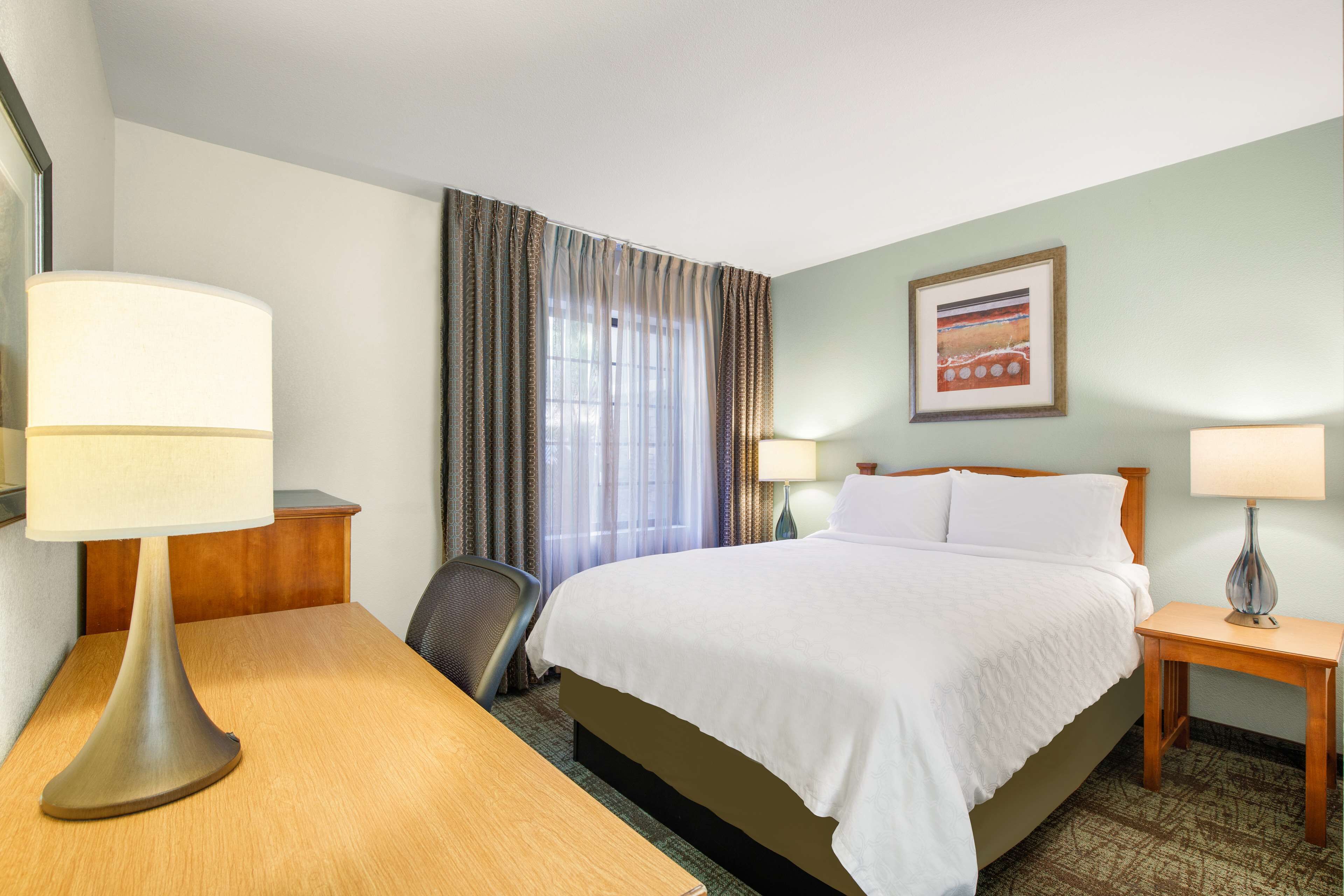 A hotel room with a bed, two bedside lamps, a desk with a lamp, a chair, and a window with curtains.