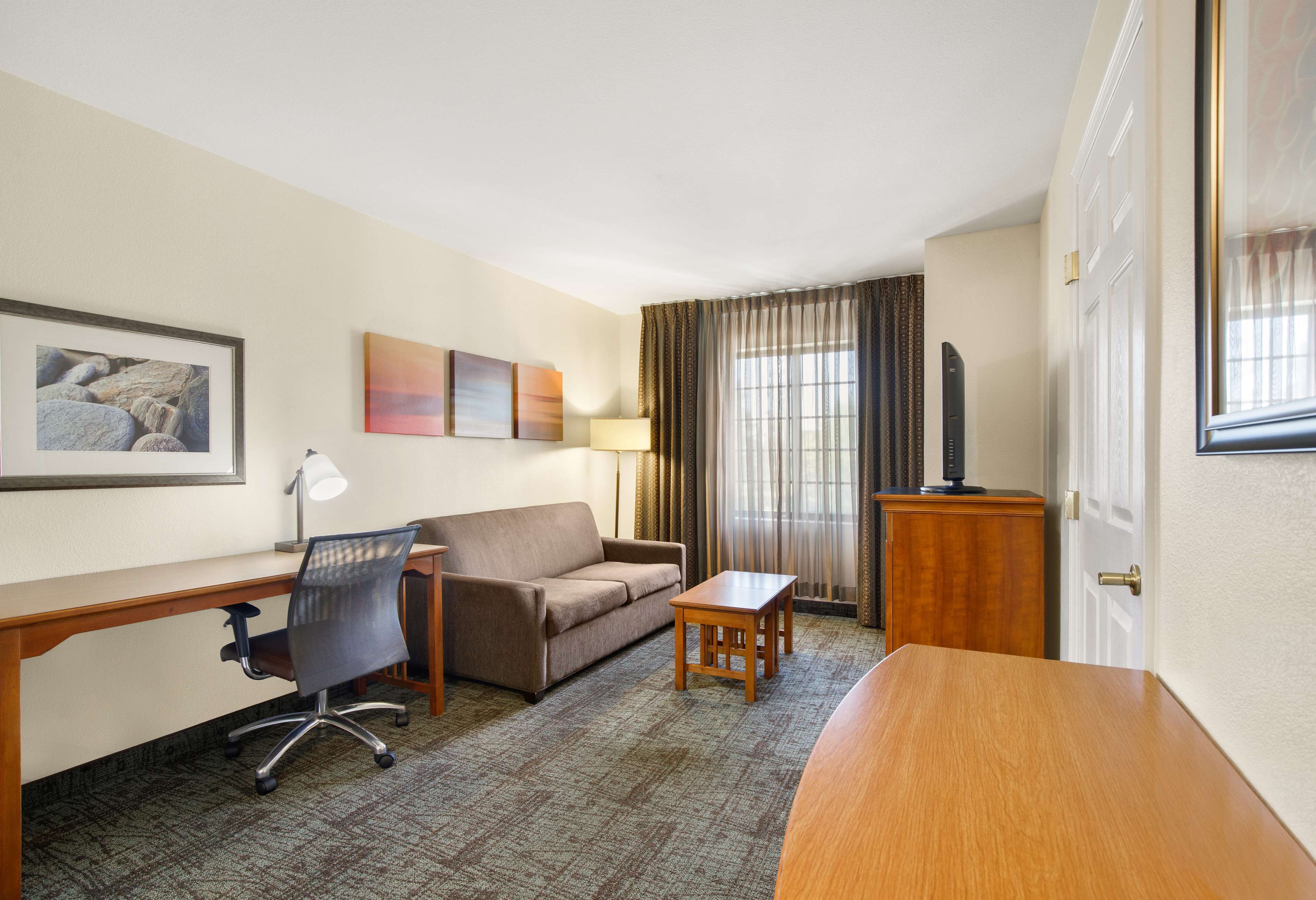 A hotel room with a wooden desk, a brown sofa, a small wooden table, and a TV on a wooden stand.