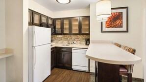 A compact kitchen with dark cabinets, a white refrigerator, and a dishwasher. There is a small dining area with a table and chairs. - Sonesta ES Suites San Diego - Rancho Bernardo (San Diego)