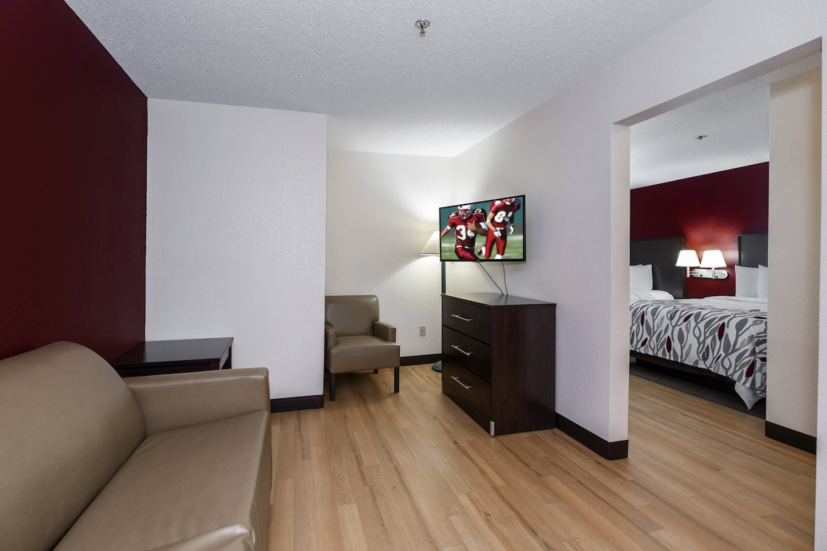 A hotel room with a sofa, a TV mounted on the wall, a desk, a chair, a dresser, and a bed with a red headboard.