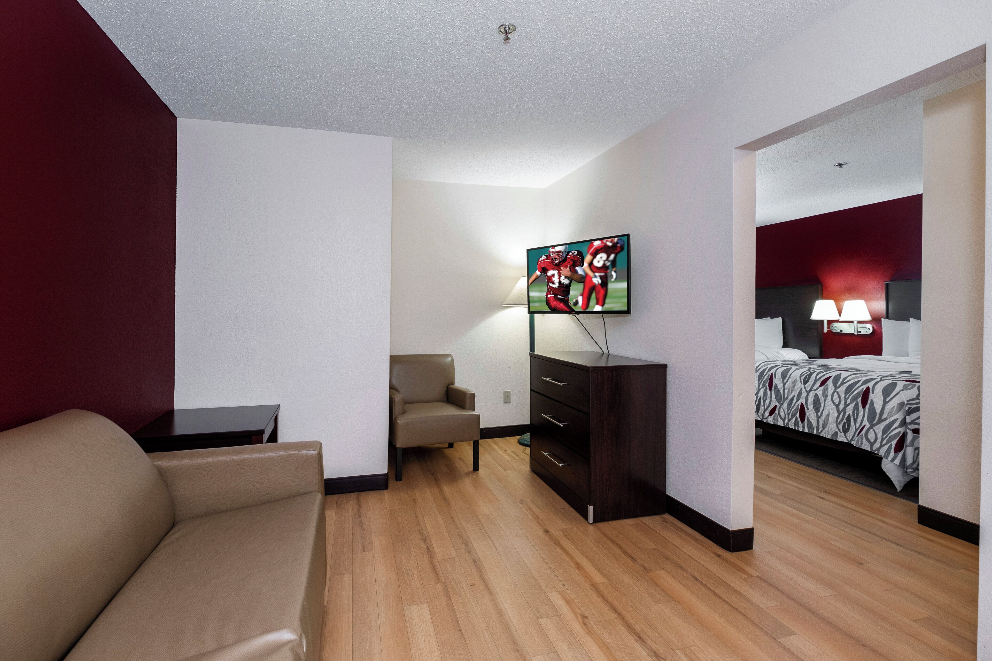 A hotel room with a sofa, a TV mounted on the wall, a desk, a chair, a wardrobe, and a bed with a patterned bedspread.