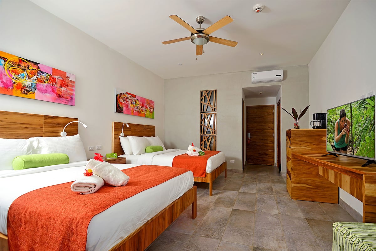 A hotel room with two beds, a TV, a ceiling fan, and artwork on the walls.
