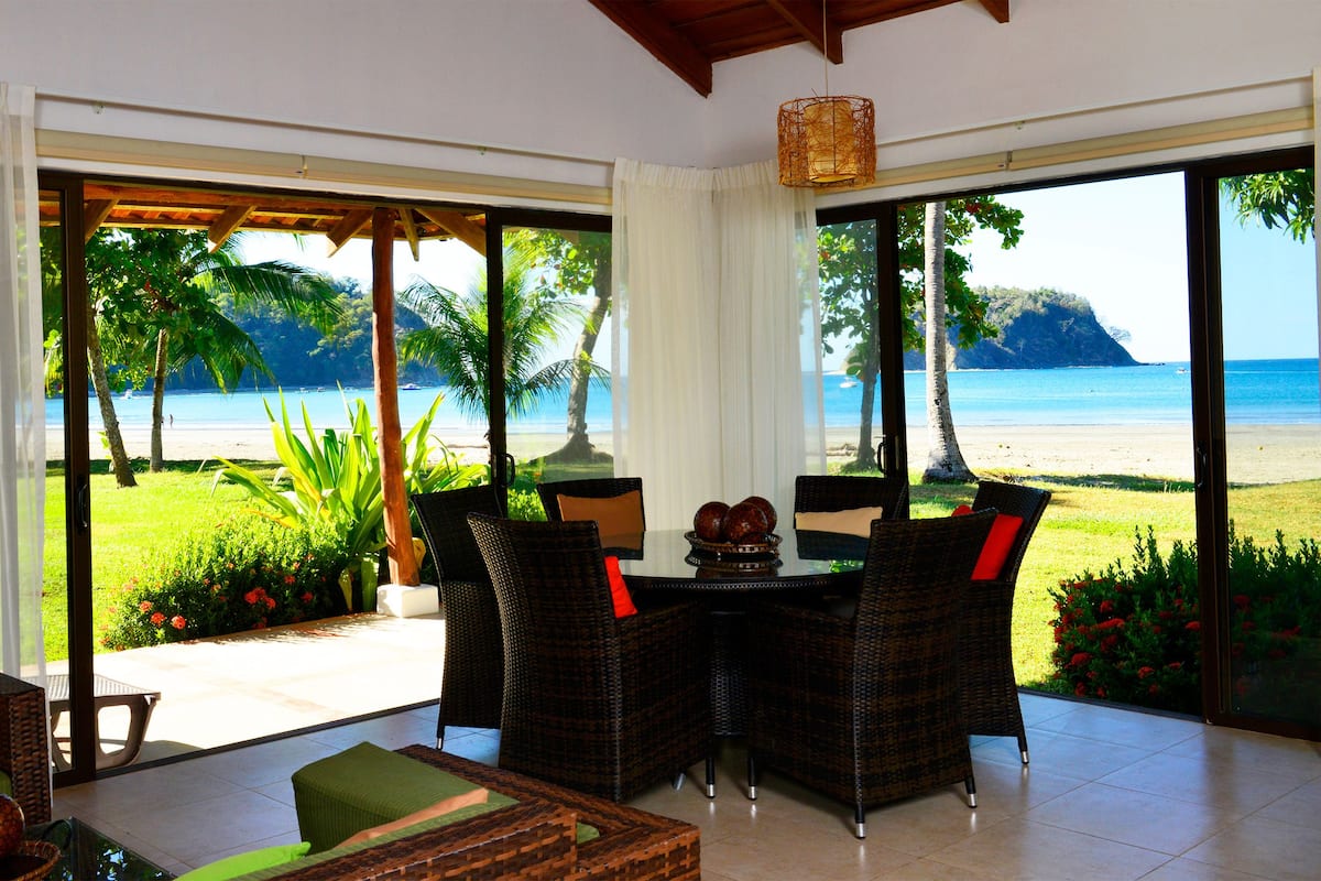 A room with a view of a beach, wicker furniture, and a table with decorative items.