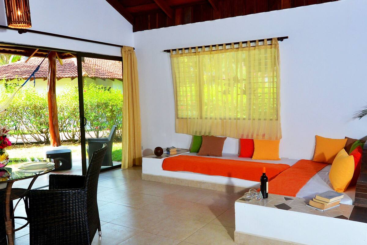 A room with a sofa, a dining table, wicker chairs, a window with yellow curtains, and a view of greenery outside.