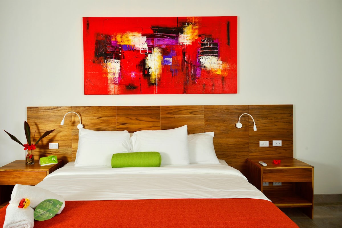 A hotel room with a wooden headboard, white bedding, a red throw blanket, and a vibrant abstract painting above the bed.