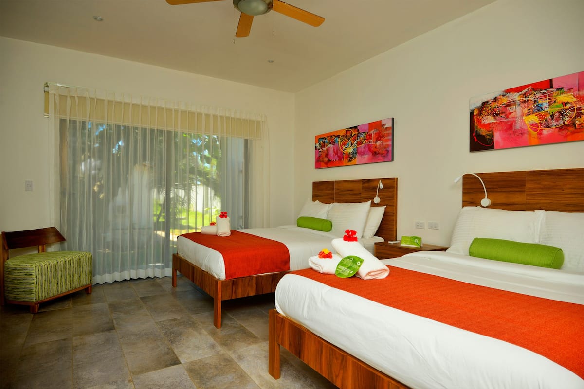 A hotel room with two beds, a wooden headboard, a green bench, and a large window with sheer curtains.