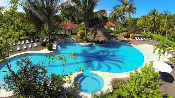 Una piscina de resort con sillas de descanso, palmeras y una cabaña de techo de paja.