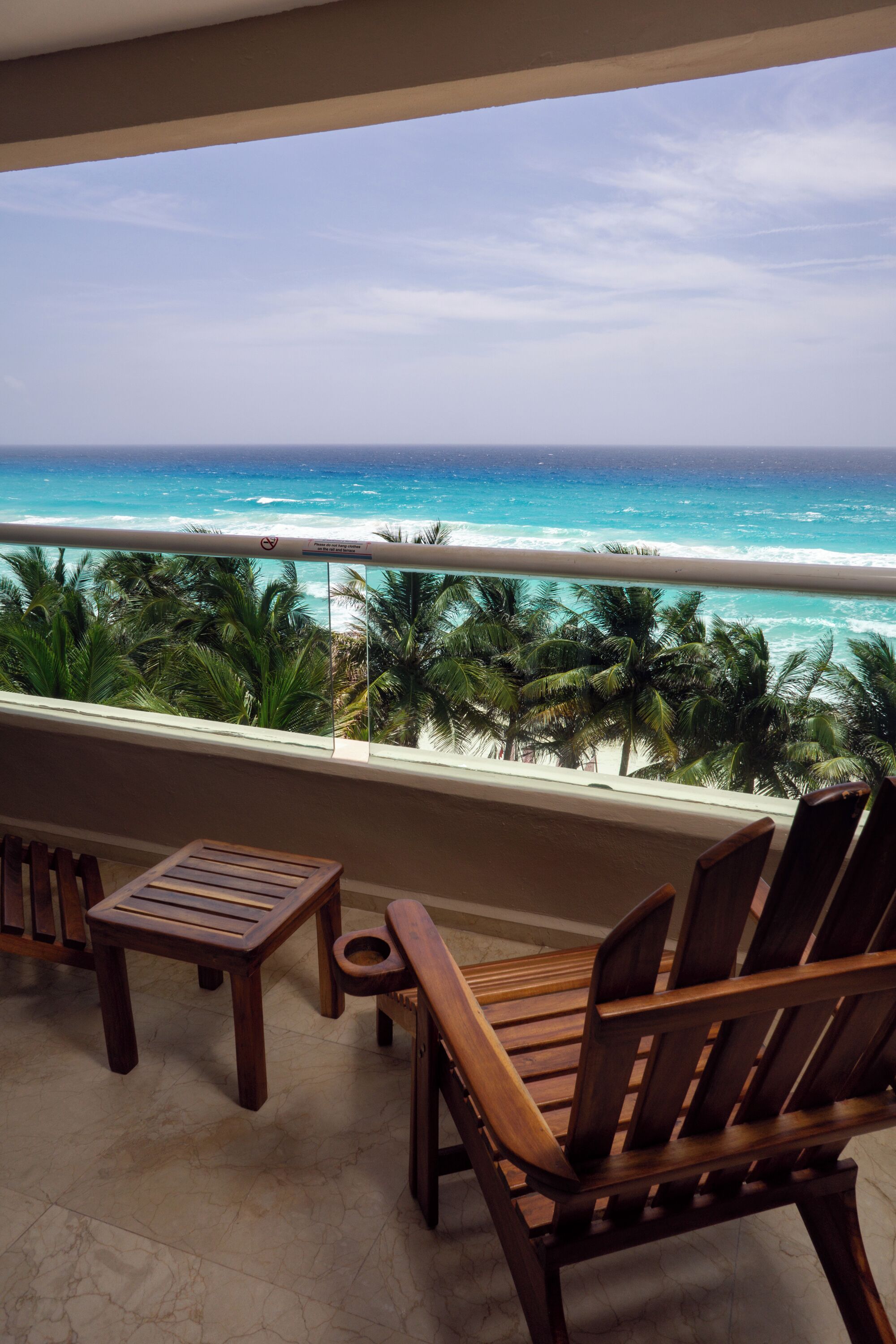 Un balcon avec des chaises en bois et une petite table, donnant sur une plage bordée de palmiers et sur l’océan.