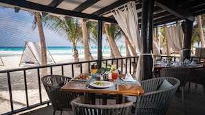 Un espace repas offrant une vue sur la plage, avec une table en bois, des chaises en osier et une rambarde en verre.