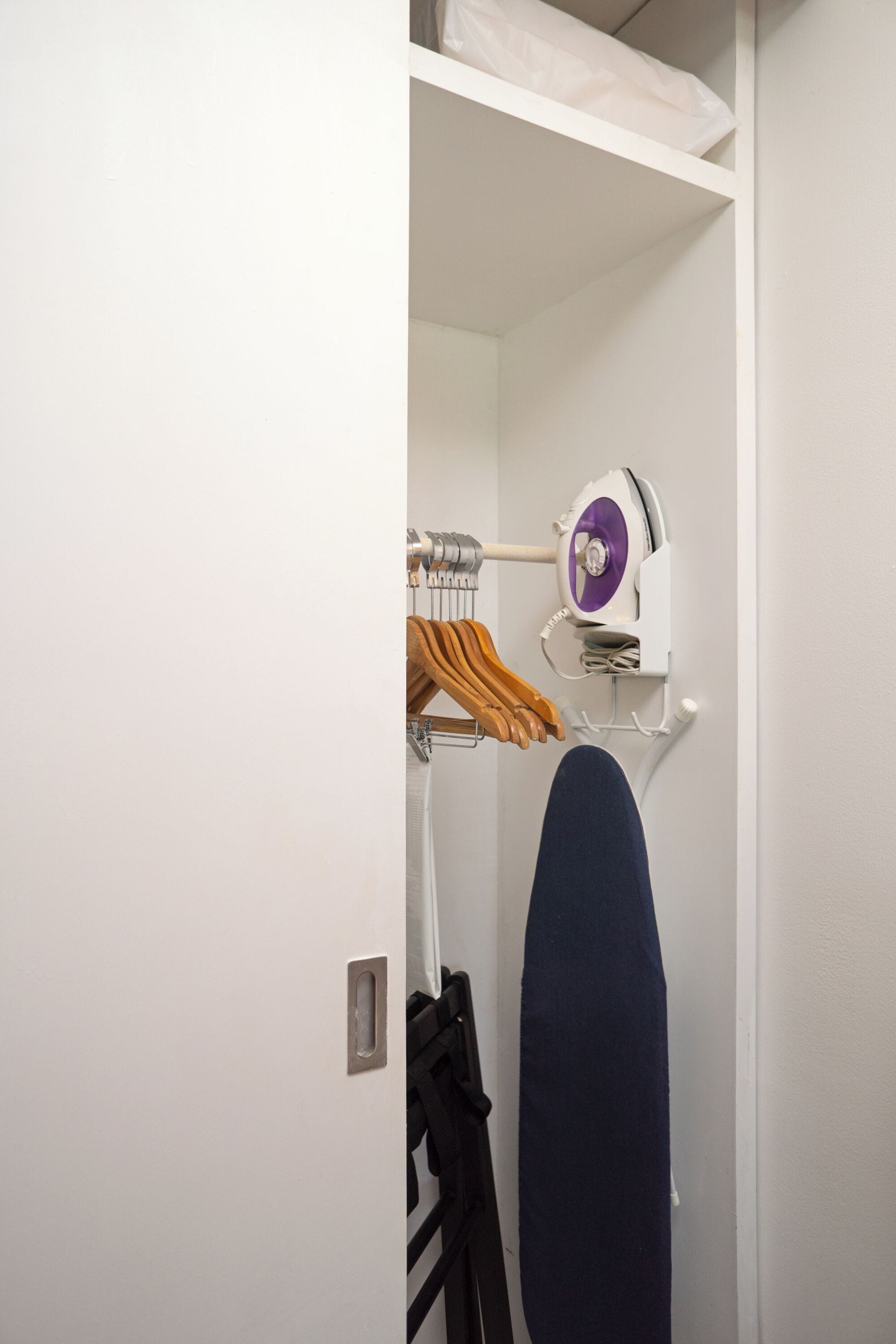 Pared blanca con tabla de planchar empotrada, un perchero con ropa y un secador de pelo fijado a la pared.