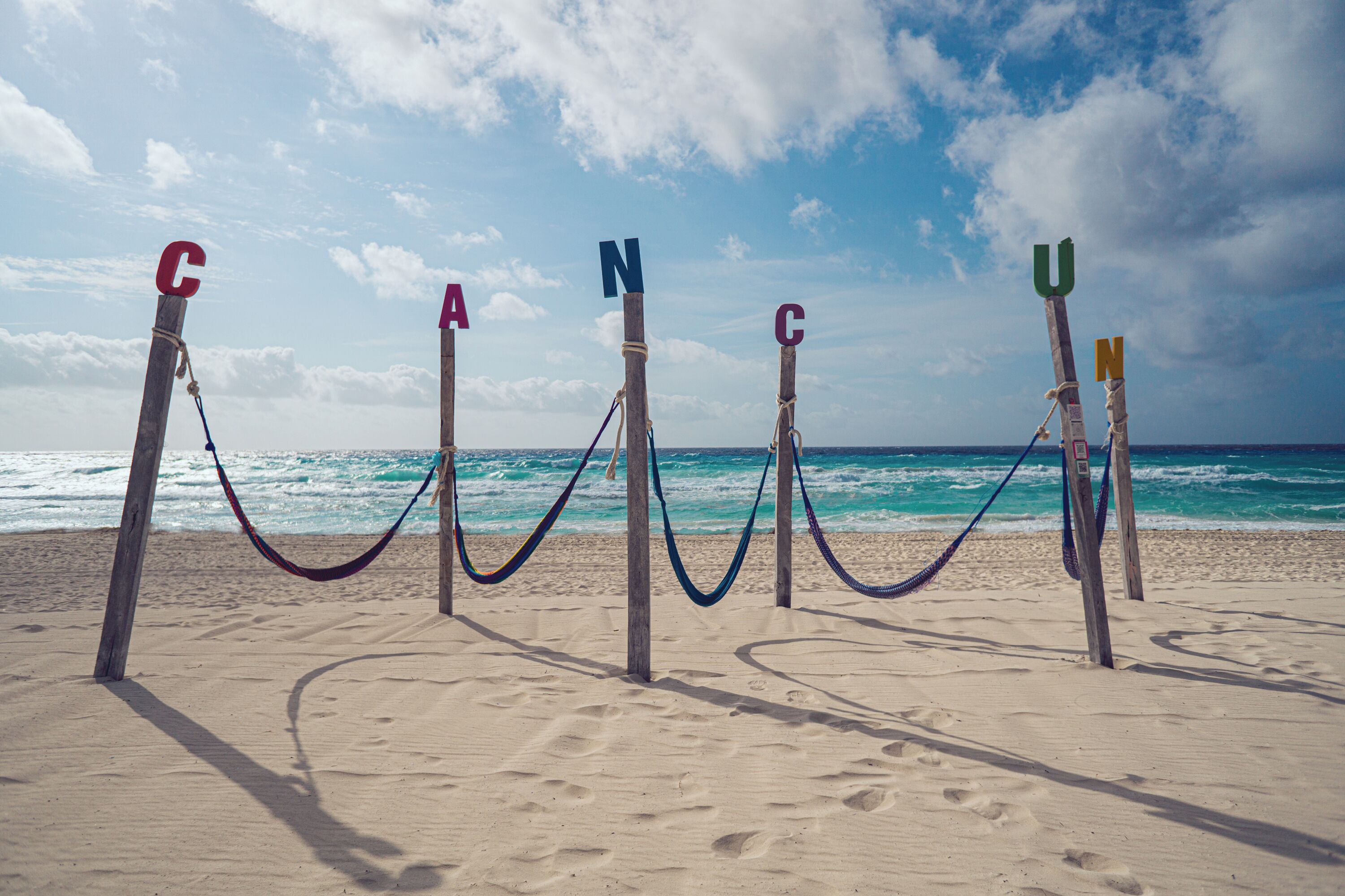 A beach with a hammock tied between wooden posts marked with letters C, A, N, C, U, and N.
