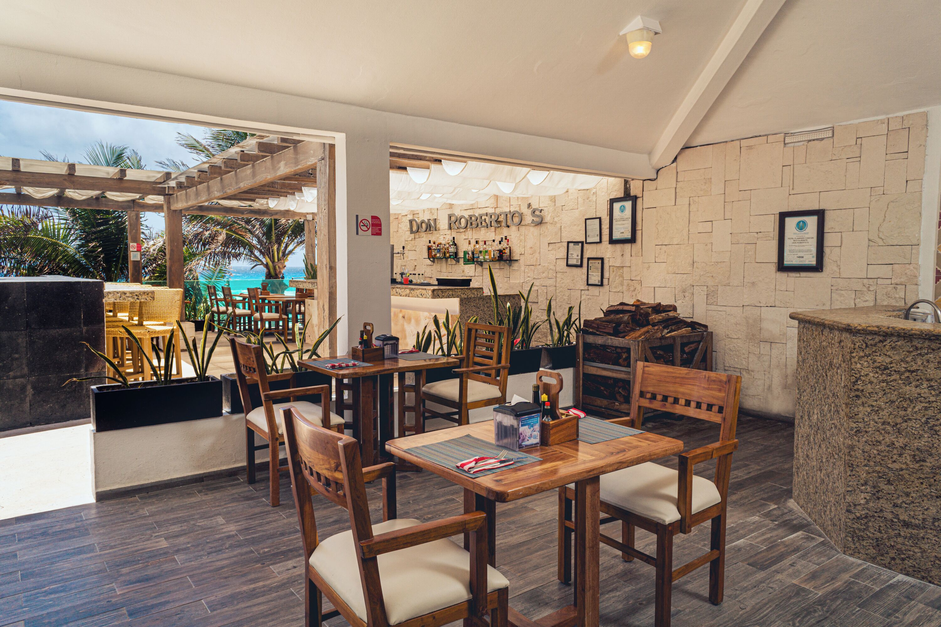 Un restaurante con mesas y sillas de madera, una pared de piedra con cuadros enmarcados y vista al océano.