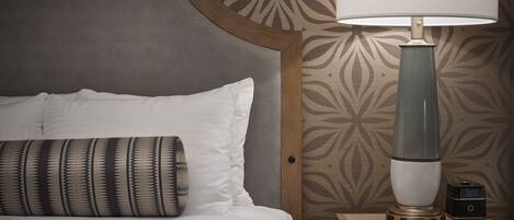 A neatly made bed with white linens and a patterned pillow, a bedside lamp, and a digital clock on a wooden bedside table.