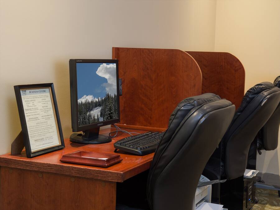 A wooden desk with a computer monitor, keyboard, and framed certificate. There are two black office chairs and a small desk lamp.