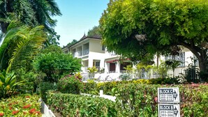 Área residencial com uma casa branca, vegetação exuberante e uma placa indicando os blocos C e B com números de apartamentos.