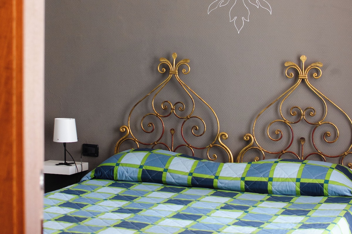 Un lit recouvert d’une couette à carreaux bleus et verts, une lampe suspendue blanche et un mur orné d’un motif en forme de fleur de lys.