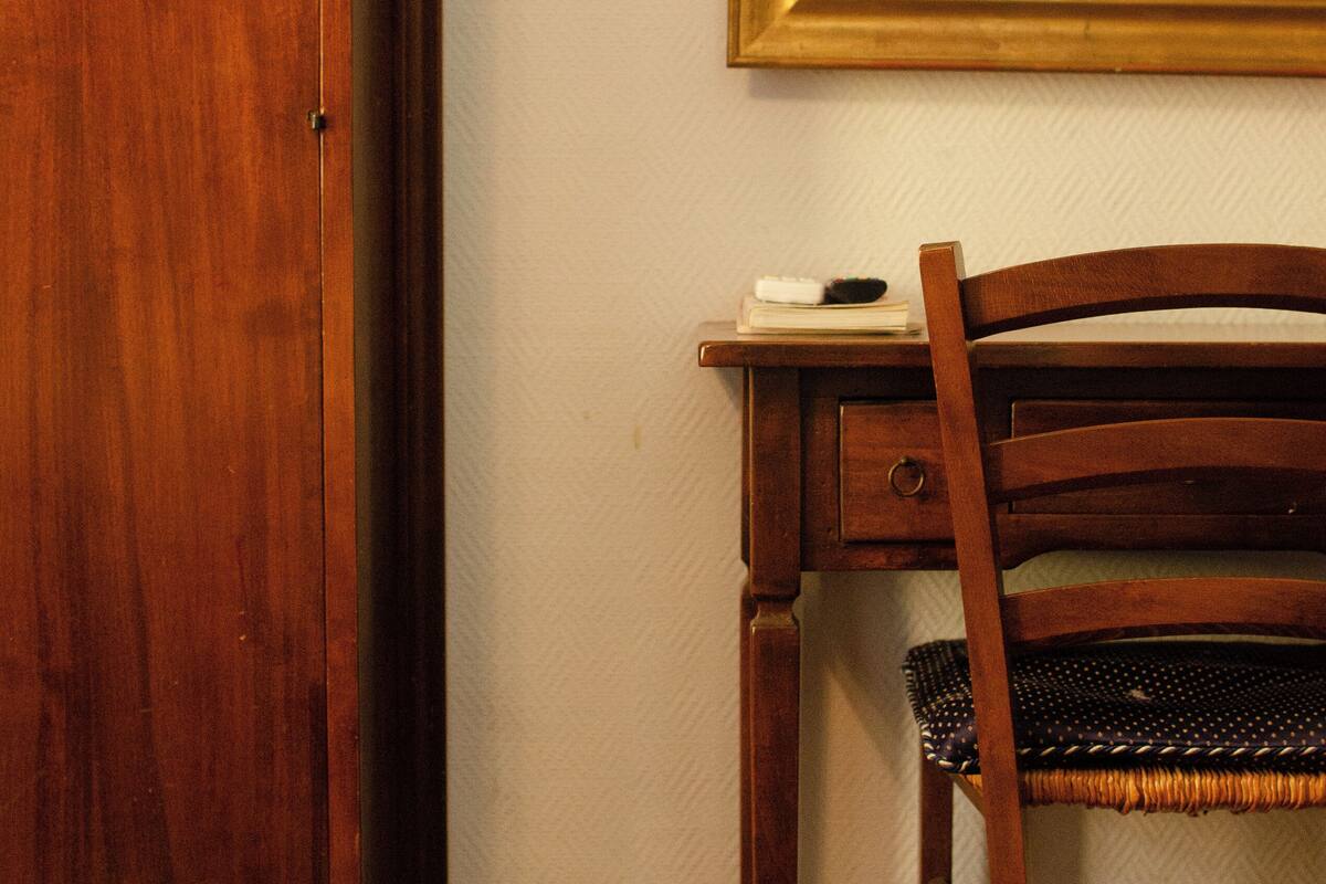 Un bureau en bois avec une chaise, un miroir et une petite table sur laquelle se trouvent un livre et une télécommande.