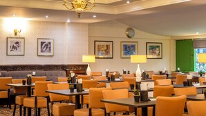Ein Essbereich eines Hotels mit orangefarbenen Stühlen und Tischen, einer Kristallleuchter, gerahmten Kunstwerken an den Wänden und einer grünen Tür im Hintergrund.