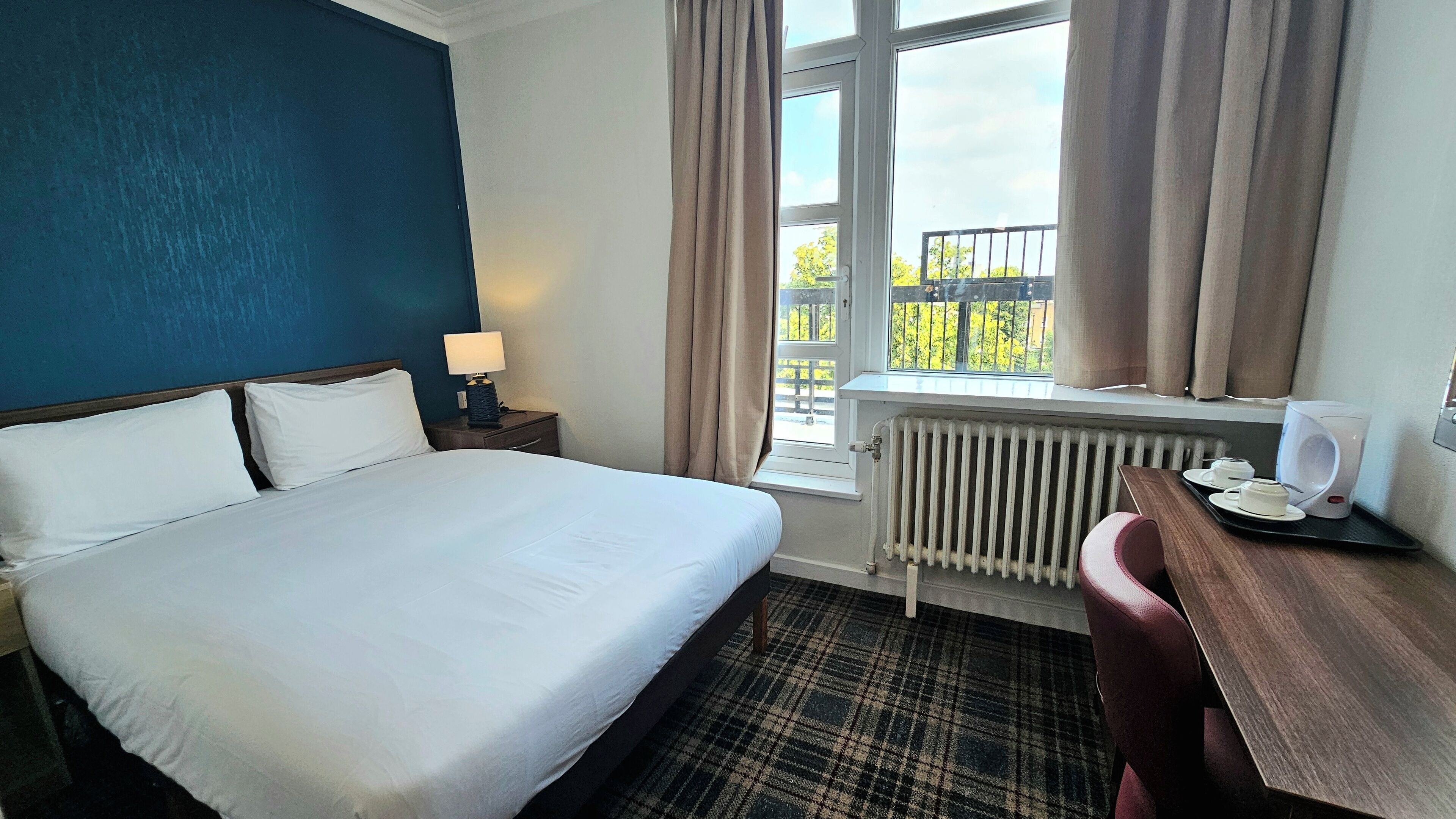 A hotel room with a bed, a desk with a coffee maker, a window with curtains, and a radiator.