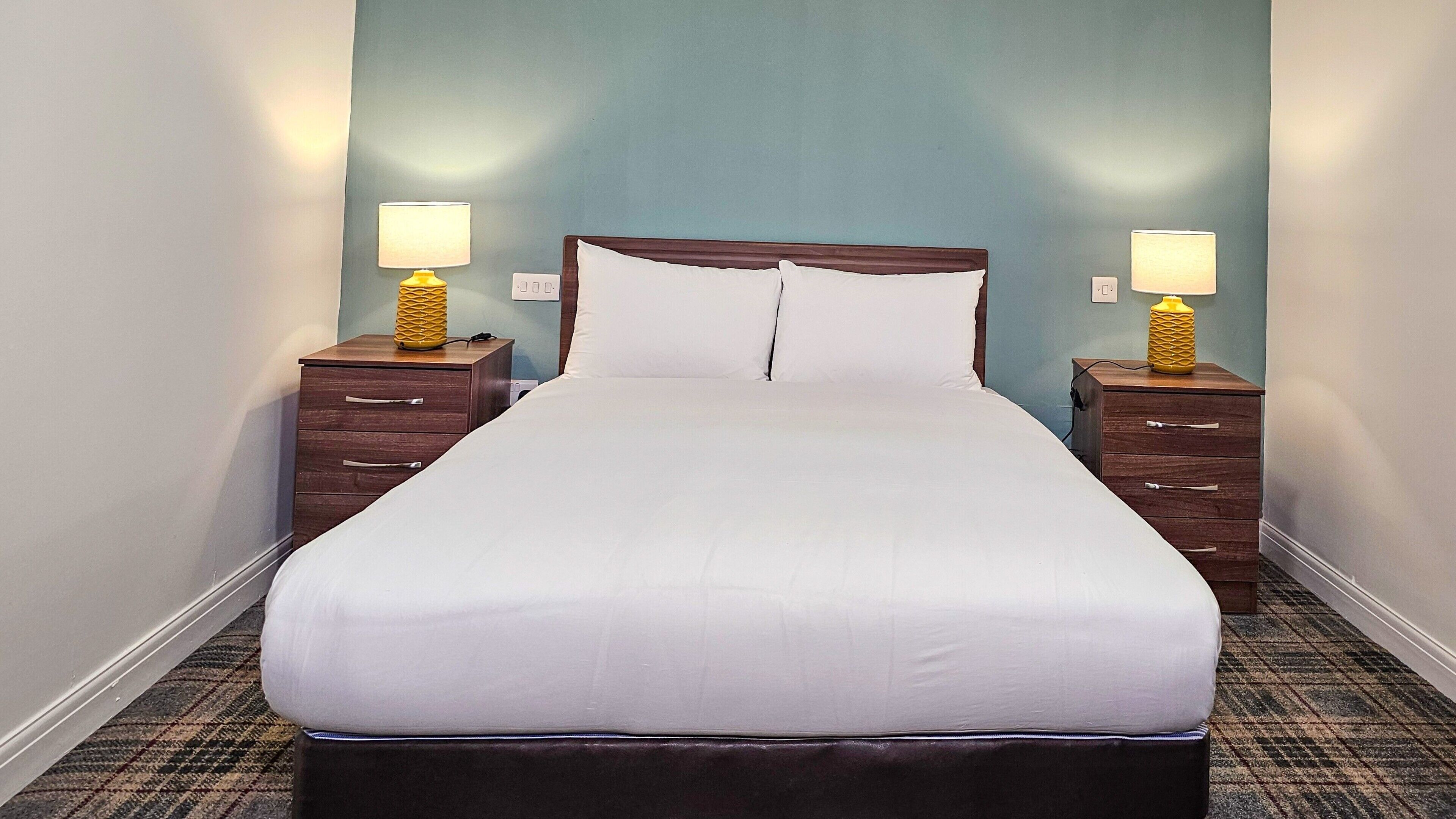 A neatly made bed with white linens, flanked by two wooden nightstands with lamps, in a room with teal walls and a carpeted floor.