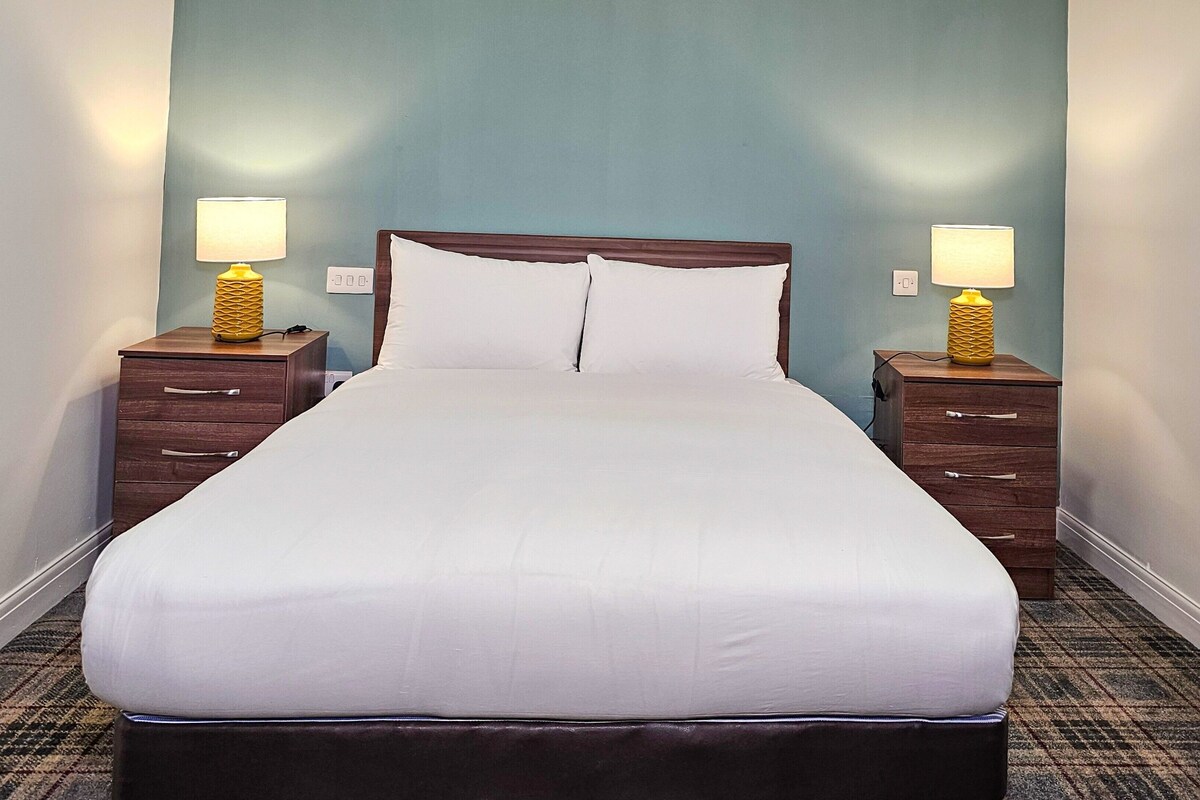 A neatly made bed with white linens, flanked by two wooden nightstands with lamps, in a room with teal walls and a carpeted floor.