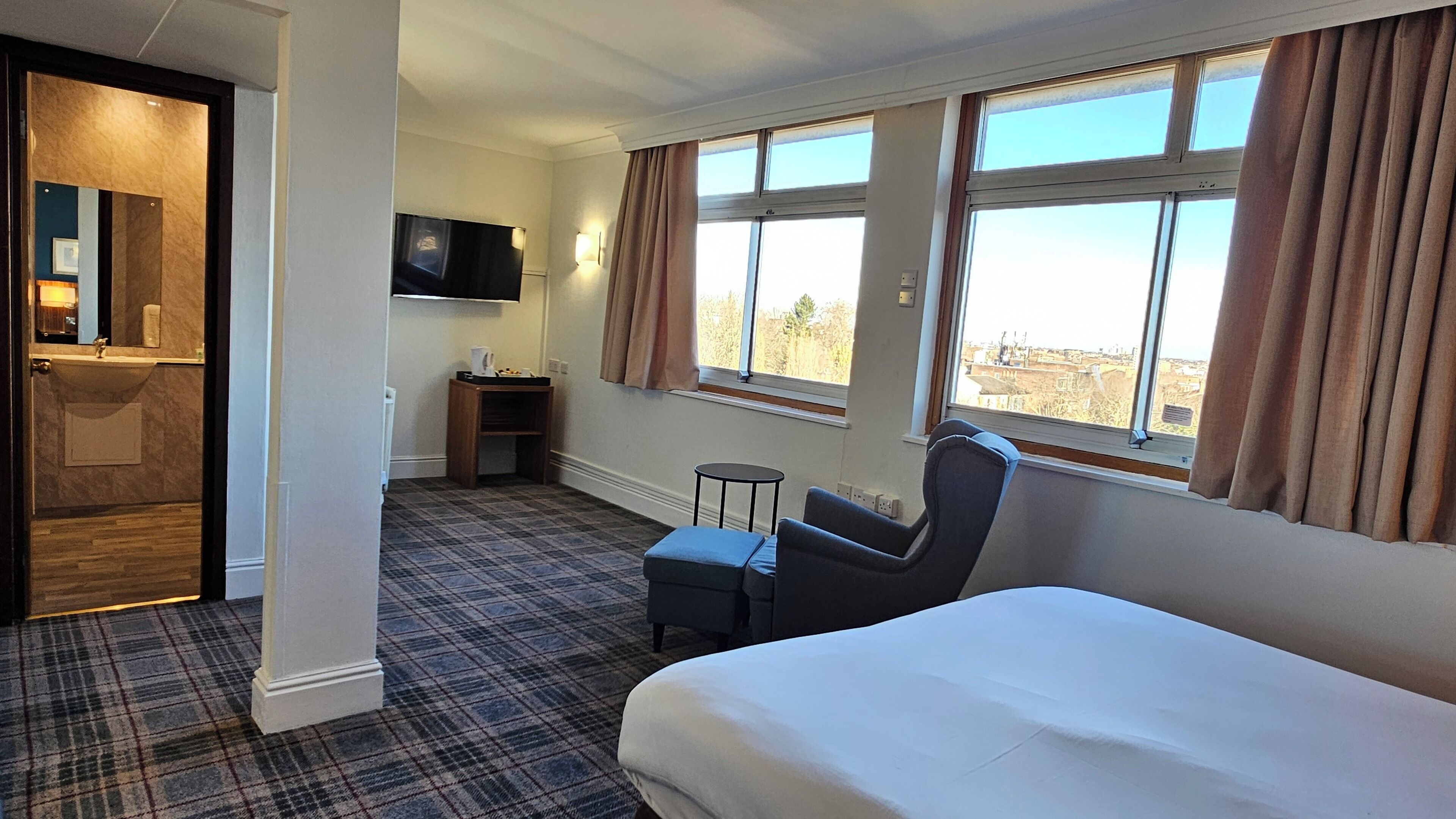 A hotel room with a bed, a chair, a small table, a TV, and a window with curtains.