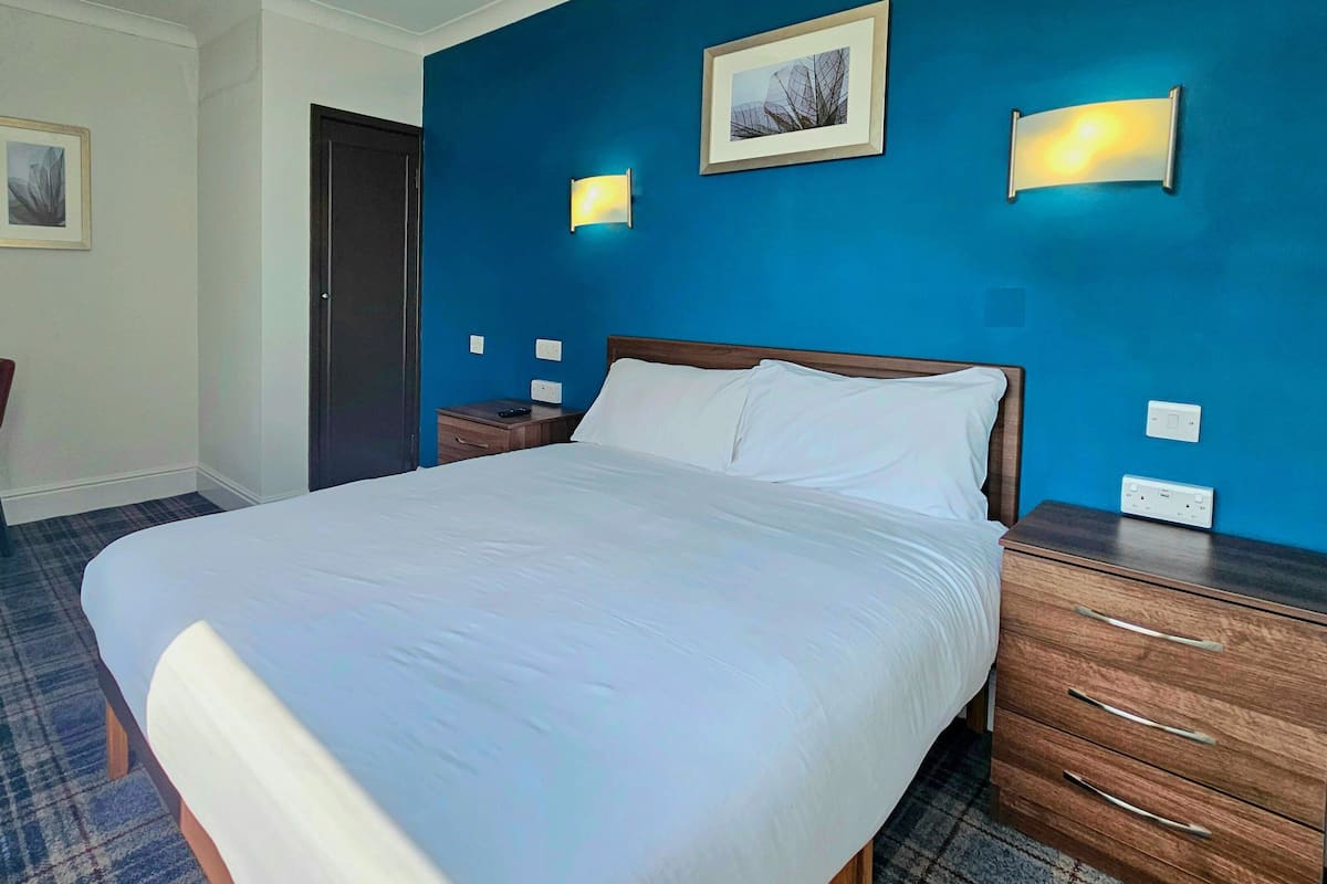 A hotel room with a large bed, a desk with a chair, and two wall-mounted lights.