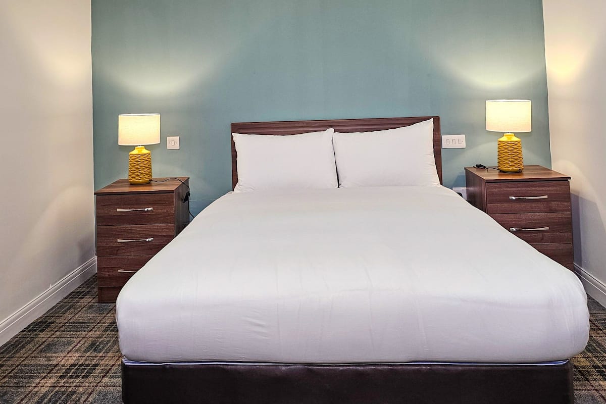 A neatly made bed in a hotel room with a wooden headboard, two bedside tables with lamps, and a carpeted floor.
