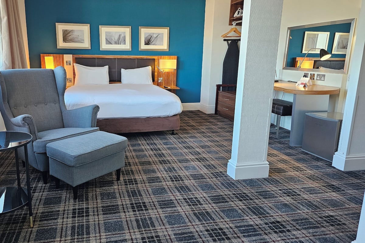 A hotel room with a bed, a grey armchair, a small table, and a desk with a lamp.
