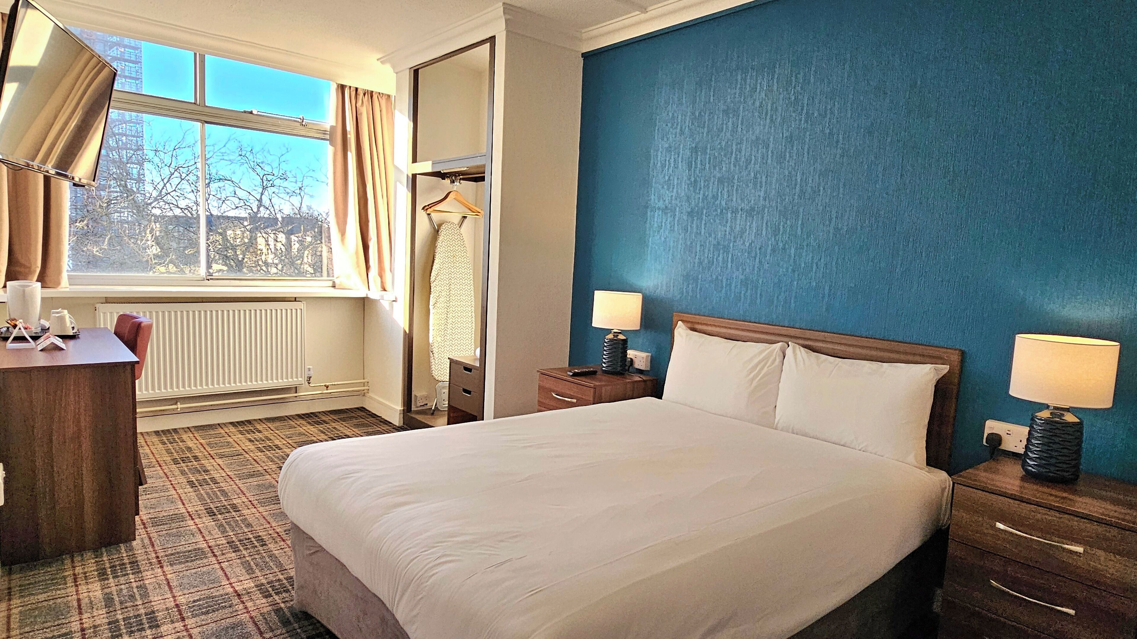 A hotel room with a large bed, bedside tables with lamps, a dresser, a desk with a lamp, a window with curtains, and a radiator.