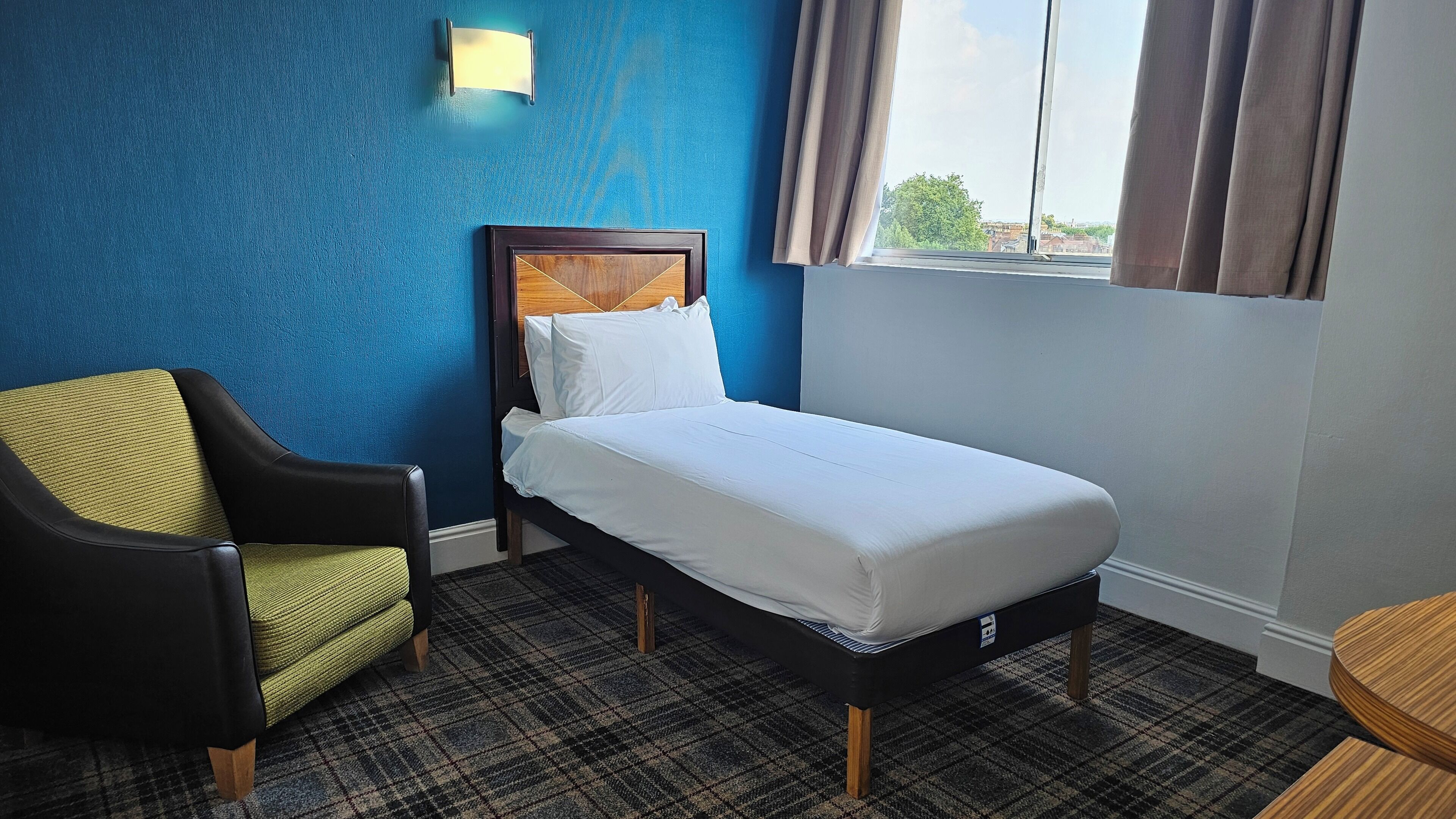 Une chambre d’hôtel avec un lit simple, une chaise, une petite table et une fenêtre avec des rideaux.