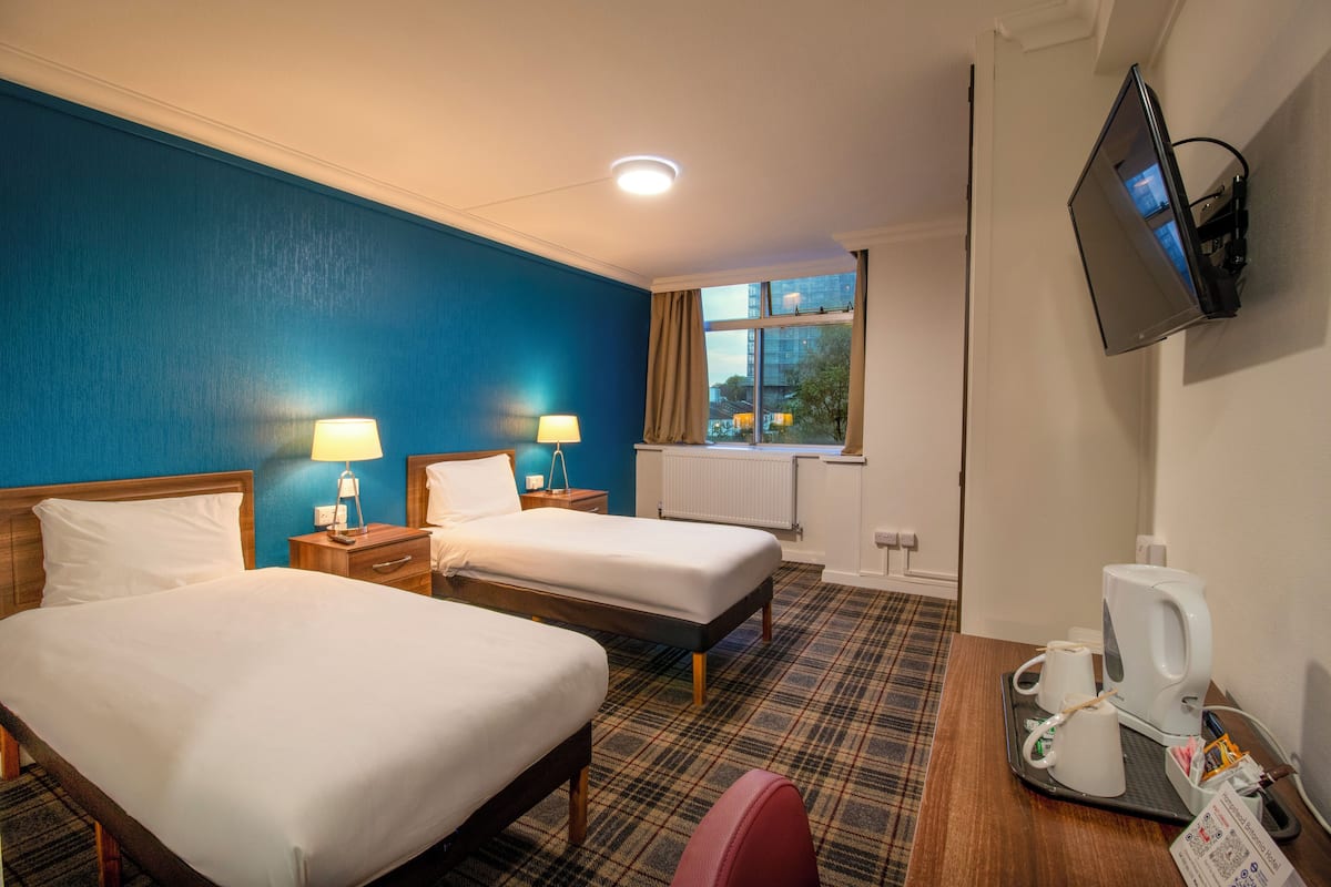 A hotel room with two beds, a television mounted on the wall, a coffee maker, and a window with a view of buildings outside.