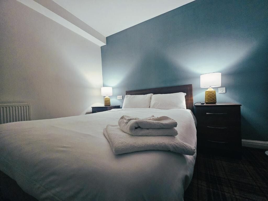 A hotel room with a neatly made bed, two bedside lamps, a dark wooden nightstand, and a blue accent wall.