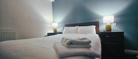 A hotel room with a neatly made bed, two bedside lamps, a dark wooden nightstand, and a blue accent wall.