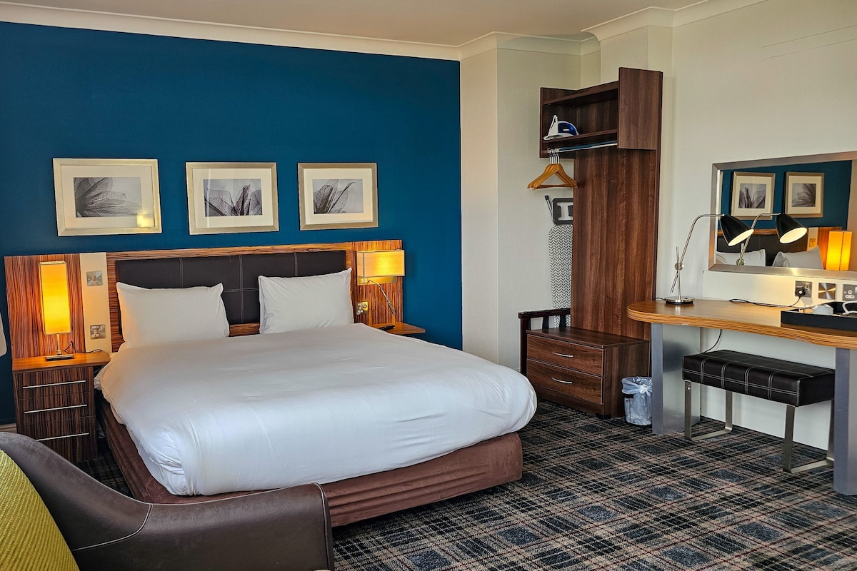 A hotel room with a large bed, a desk with a chair, a wardrobe, and three framed pictures on the wall.