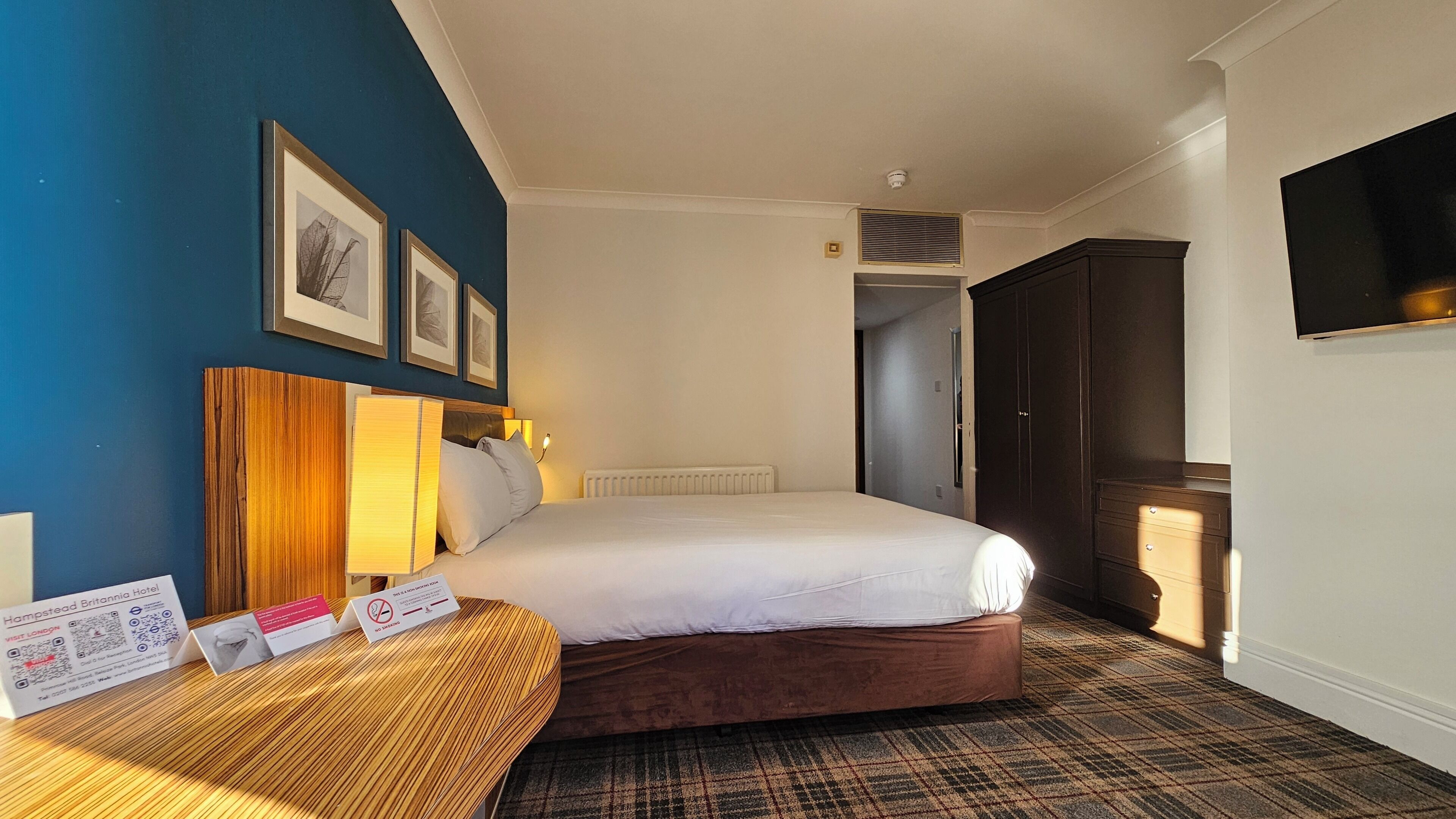 A hotel room with a bed, a bedside table with a lamp, a wall-mounted TV, and framed pictures.
