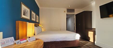 A hotel room with a bed, a bedside table with a lamp, a wall-mounted TV, and framed pictures.